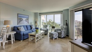 Una sala de estar con un sofá azul, una mesita de centro, sillones estampados y vista al mar a través de amplias ventanas.