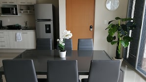 Een moderne keuken met een koelkast, magnetron en spoelbak. Er staat een eettafel met stoelen en een potorkidee. Op de muur is een houten deur en een klok te zien.