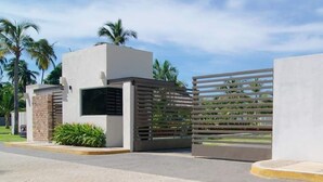 Een modern gebouw met een plat dak, een kleine ingang met een metalen hek en een verhard gebied met palmbomen op de achtergrond.