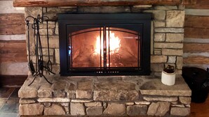 A stone fireplace with a fire, flanked by stone walls and wooden beams. - Quaint, Romantic Mountain Antique Log Cabin near Asheville! (Fairview)