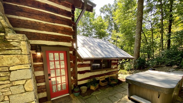 A rustic cabin with a stone exterior, a red door, and a hot tub on a stone deck. - Quaint, Romantic Mountain Antique Log Cabin near Asheville! (Fairview)