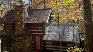 A rustic cabin with a stone chimney, a red door, and a metal roof, surrounded by autumn foliage. - Quaint, Romantic Mountain Antique Log Cabin near Asheville! (Fairview)