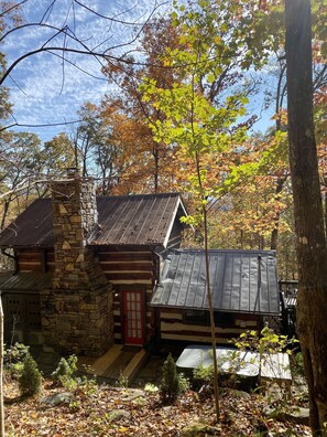 A rustic cabin with a stone chimney, a red door, and a metal roof, surrounded by autumn foliage. - Quaint, Romantic Mountain Antique Log Cabin near Asheville! (Fairview)