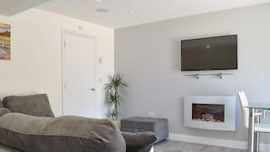 Una sala de estar moderna con un sofá gris, un televisor de pantalla plana sobre la chimenea, y una pequeña mesa con sillas.