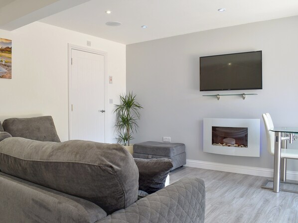 Un salon moderne avec un canapé gris, un téléviseur à écran plat fixé au-dessus d’une cheminée, et une petite table avec des chaises.
