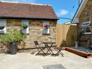 Une maison en briques avec un toit en tuiles, une clôture en bois, un jardin agrémenté d’une table et de chaises, et des jardinières suspendues.