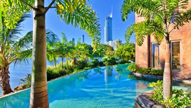 A pool with clear blue water, surrounded by lush greenery and palm trees. In the background, there is a modern building with a tall, narrow design. - Casa Albero Via Roma (Surfers Paradise)