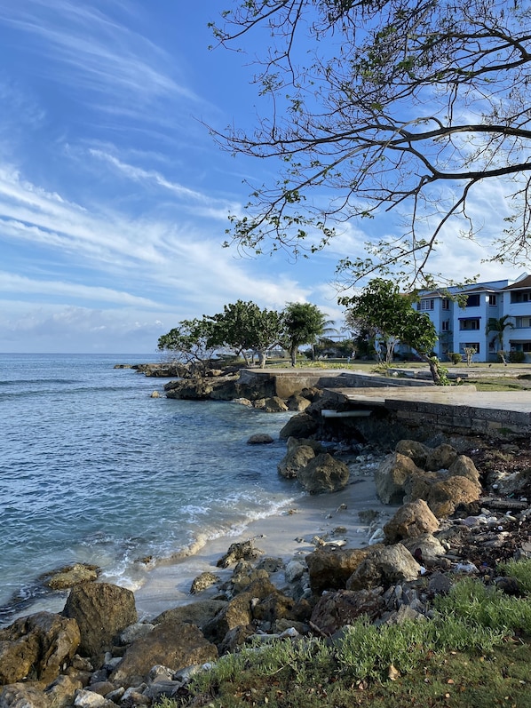 A coastal scene with rocky shoreline, clear blue sky, and a building with balconies overlooking the sea.