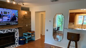 Ein modernes Wohnzimmer mit Fernseher, der ein Sportevent zeigt, einer Holzwand, einem Kamin, einem Esstisch und einer Küchenzeile mit Fenster.