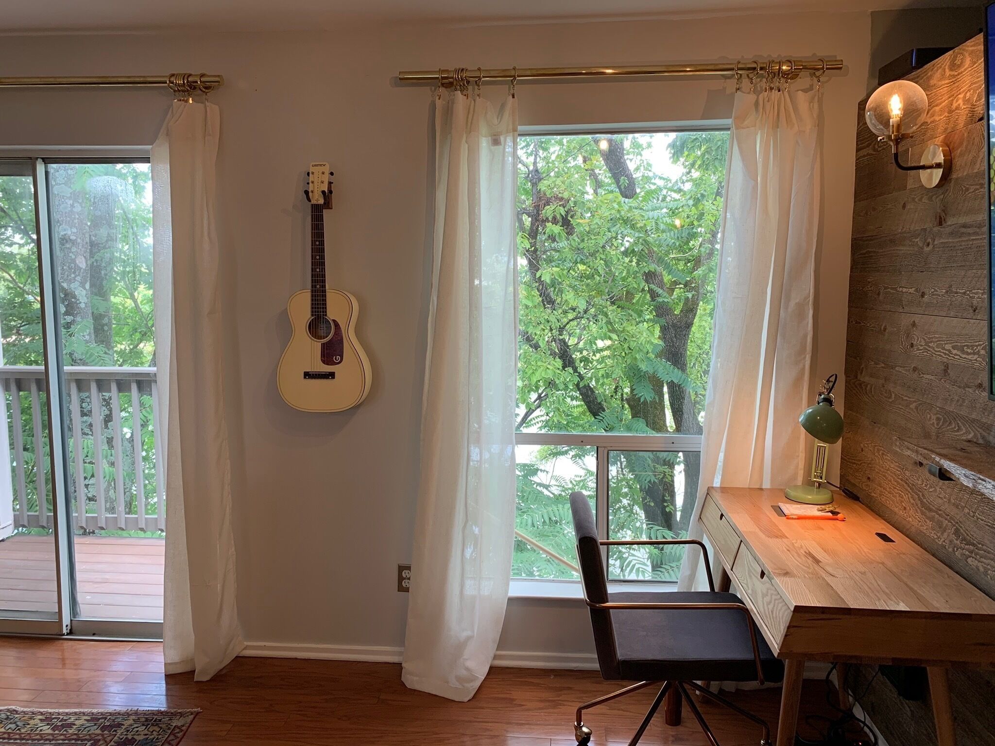 A room with a wooden desk, a chair, a guitar, and large windows with curtains.