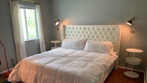 Ein Schlafzimmer mit einem gepolsterten Kopfteil, weißer Bettwäsche, zwei Nachttischen mit Lampen, einem Fenster mit Vorhängen und einem gemusterten Teppich.