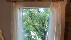 Ein Fenster mit transparenten weißen Vorhängen, eine an der Wand angelehnte Holzgitarre und Blick auf grüne Bäume draußen.
