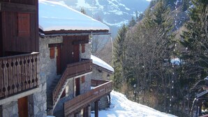 Un chalet en bois avec une façade en pierre, un balcon et un toit enneigé, entouré d’arbres et de montagnes.