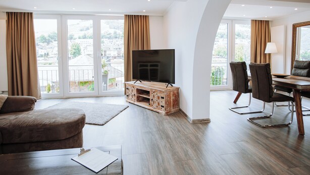 Una sala moderna con televisor de pantalla plana, mueble de madera para entretenimiento, sofá, mesa de comedor con sillas y ventanas amplias con cortinas.