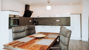 Una cocina moderna con armarios blancos, horno empotrado, cafetera y un comedor con mesa de madera y sillas acolchadas.