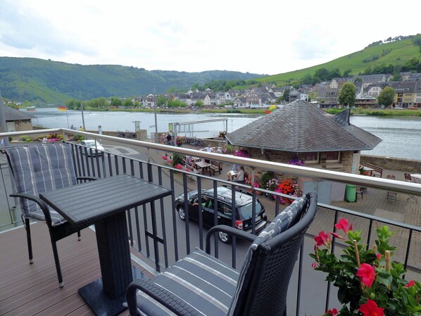 A deck with outdoor seating overlooking a river, surrounded by buildings and hills.
