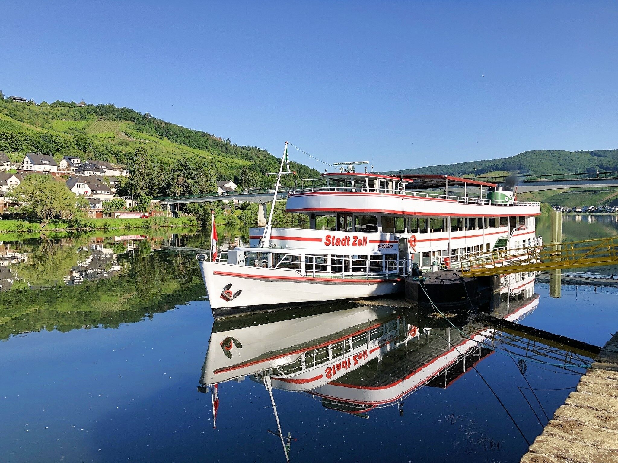 Un barco blanco y rojo llamado Stadt Zell atracado en un río tranquilo, con un puente y casas al fondo.