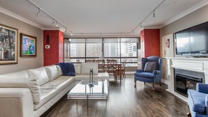 Een moderne woonkamer met een witte leren bank, een glazen salontafel en een blauwe fauteuil. Er is een open haard met een televisie aan de rechterkant en grote ramen met uitzicht op de stad.