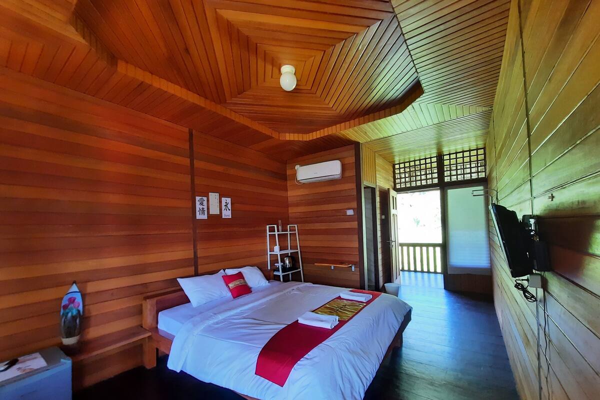 A wooden cabin-style room with a bed, a small table, a chair, a shelf, and a TV.