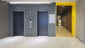A modern hotel lobby with three elevators, a digital information board, and a wooden floor.