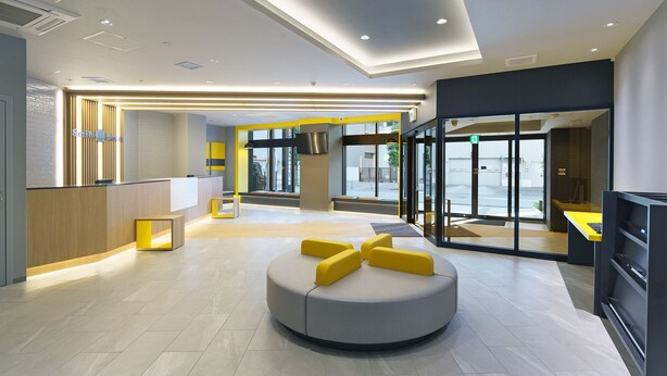 A modern hotel lobby with a reception desk, a seating area with yellow cushions, and large glass doors leading outside.