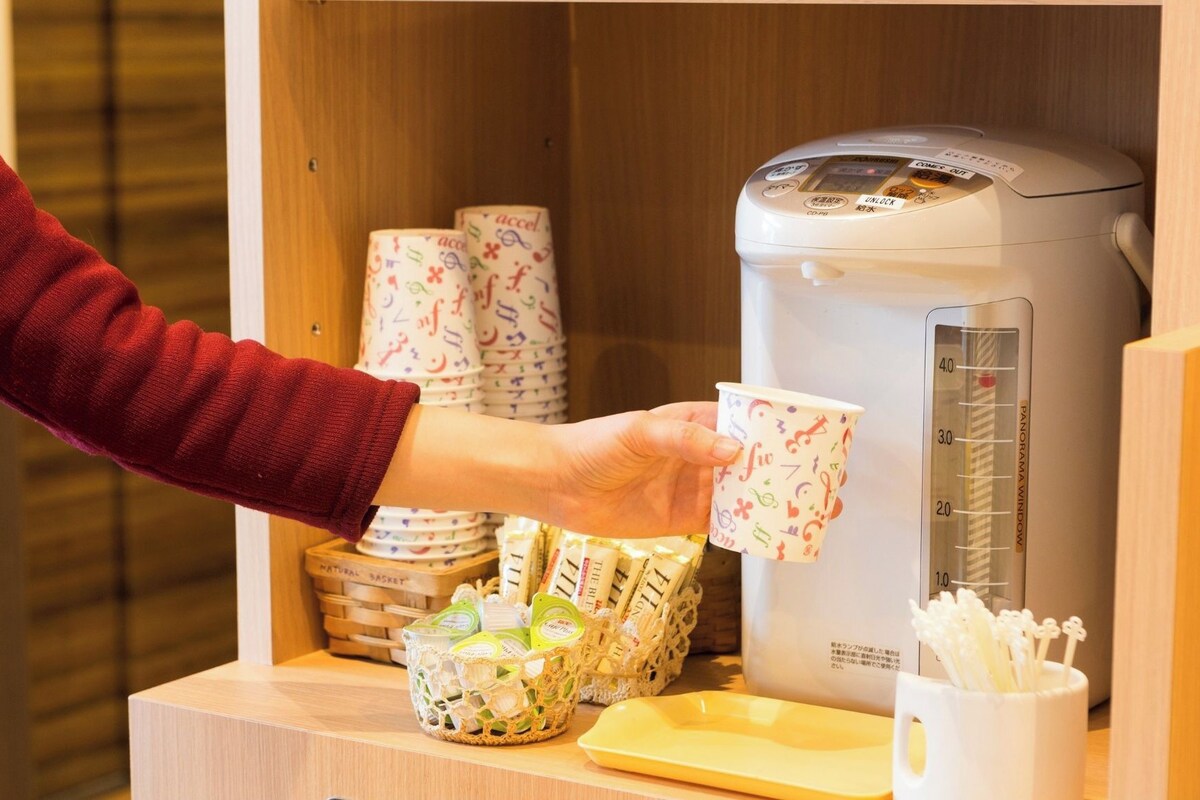 A person serving coffee from a machine, with paper cups and a basket of packaged goods on the counter.