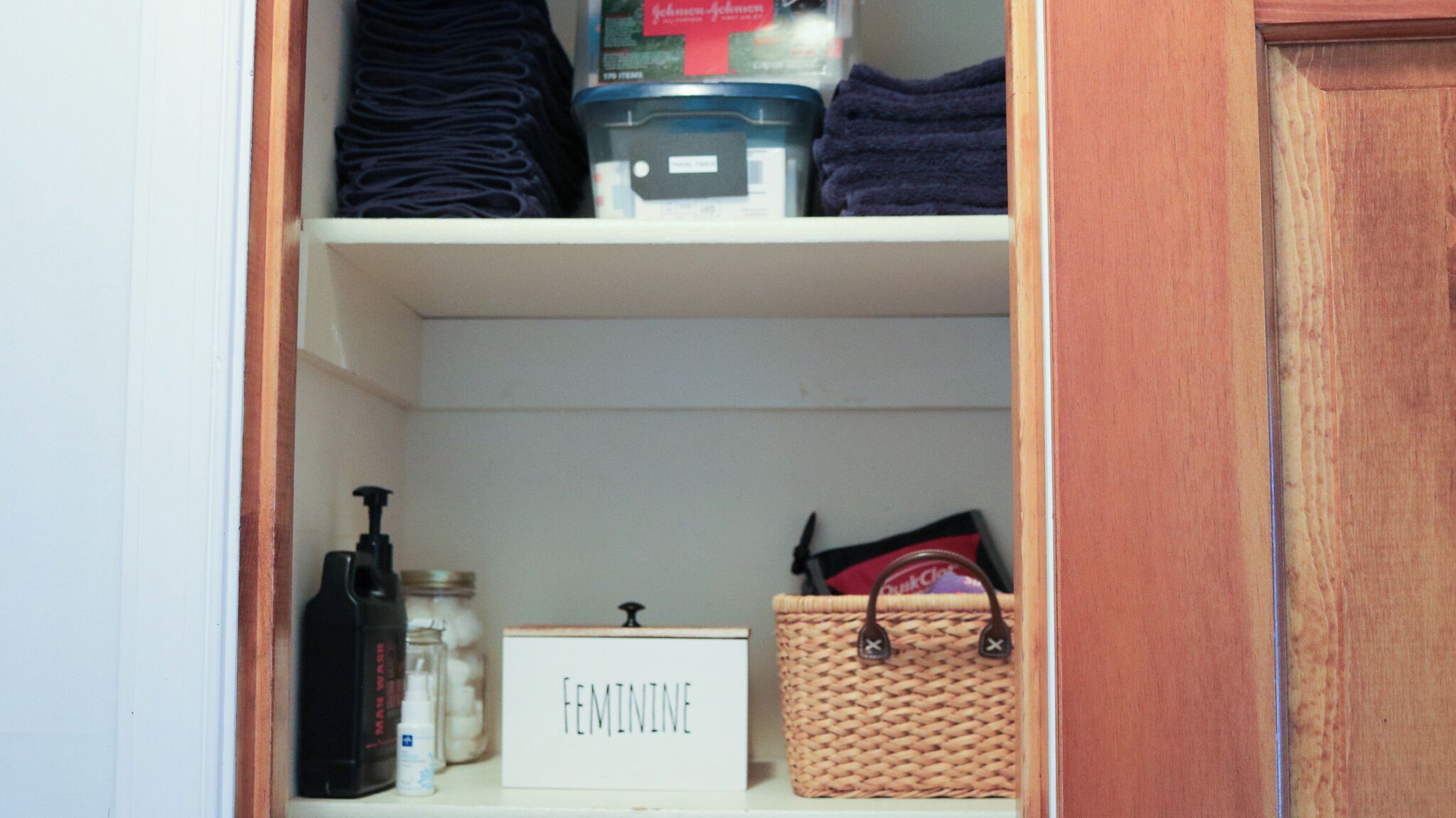 A storage area with shelves containing folded towels, a container of Johnson & Johnson baby powder, a bottle of hand soap, a jar of cotton balls, and a wicker basket with a red item.