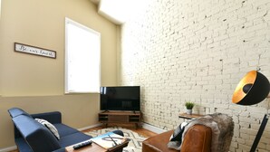 Un salon avec un canapé bleu, un fauteuil en cuir marron, un téléviseur et un mur en briques.
