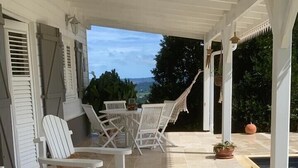 Un espacio de estar al aire libre con techo, con sillas y mesas blancas, una piscina y vistas al paisaje.