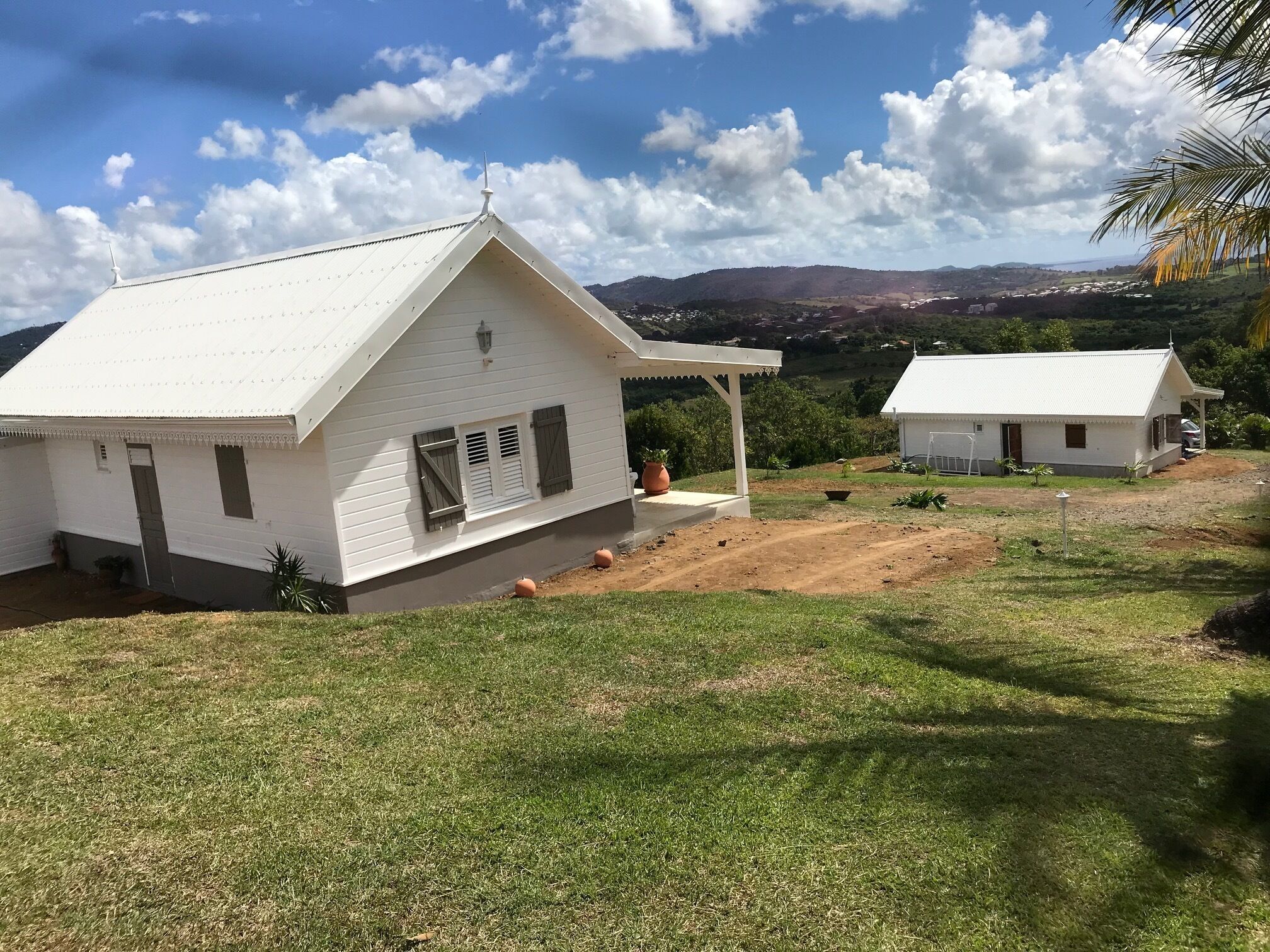 A white building with a sloped roof and a covered porch, surrounded by a grassy area and a dirt path.