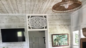 Una habitación con techo de madera, televisor montado en la pared, una silla de mimbre, una mesa de comedor con sillas, una planta en maceta, una cama con cabecera blanca y un cuadro enmarcado en la pared.
