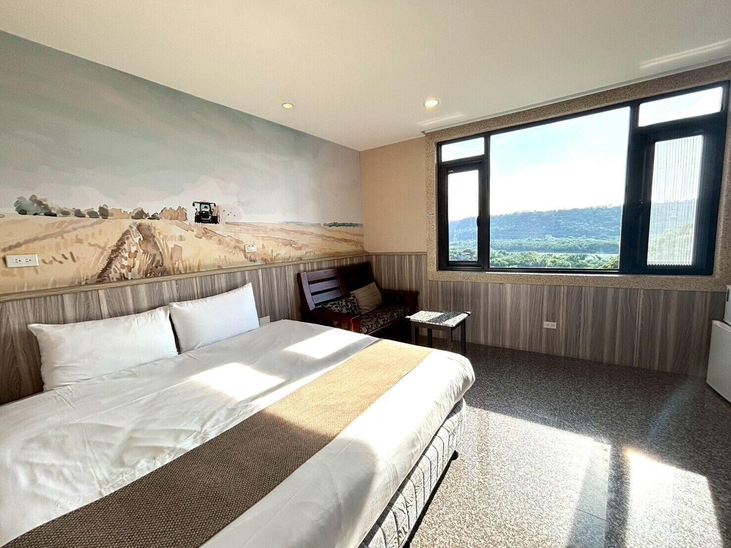 Habitación de hotel con una cama grande, una mesita pequeña, una silla y vistas a un paisaje a través de un ventanal amplio.