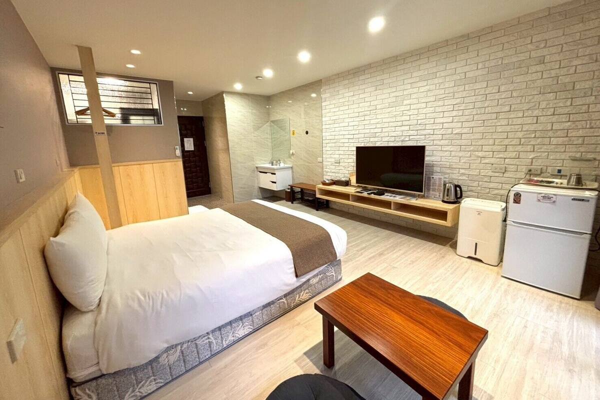 A modern hotel room with a bed, a TV, a small kitchenette, and a brick wall.