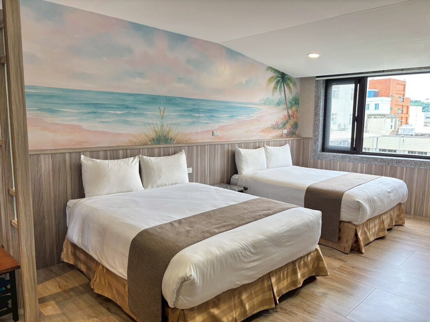 Habitació d'hotel amb dos llits, un mural de platja i una finestra amb vista a la ciutat.