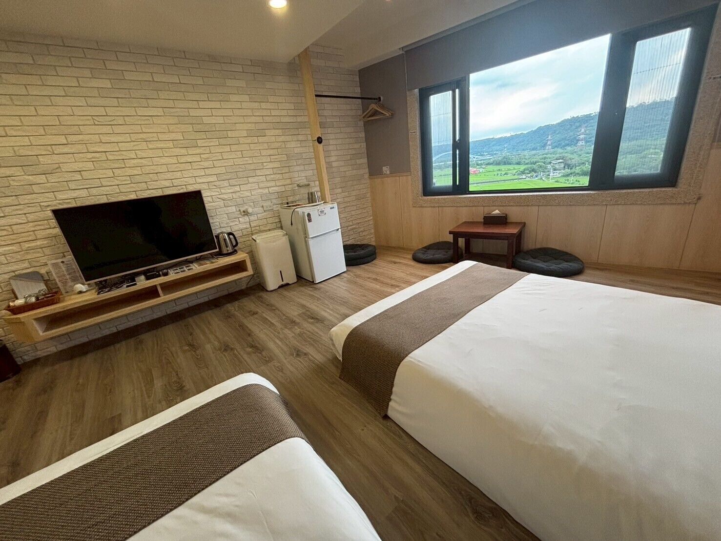 A hotel room with a brick wall, a flat-screen TV, a small fridge, a wooden table, two beds, and a large window with a view of the countryside.