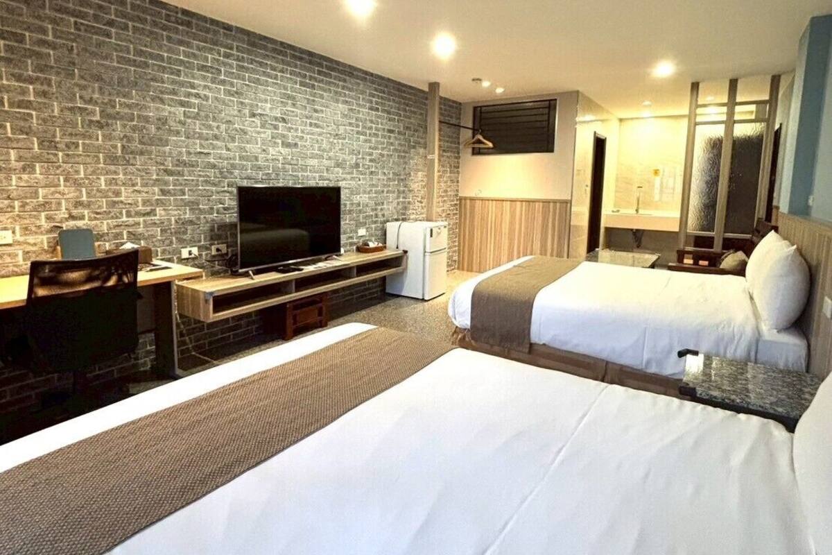 A hotel room with two beds, a TV, a desk, and a brick wall.