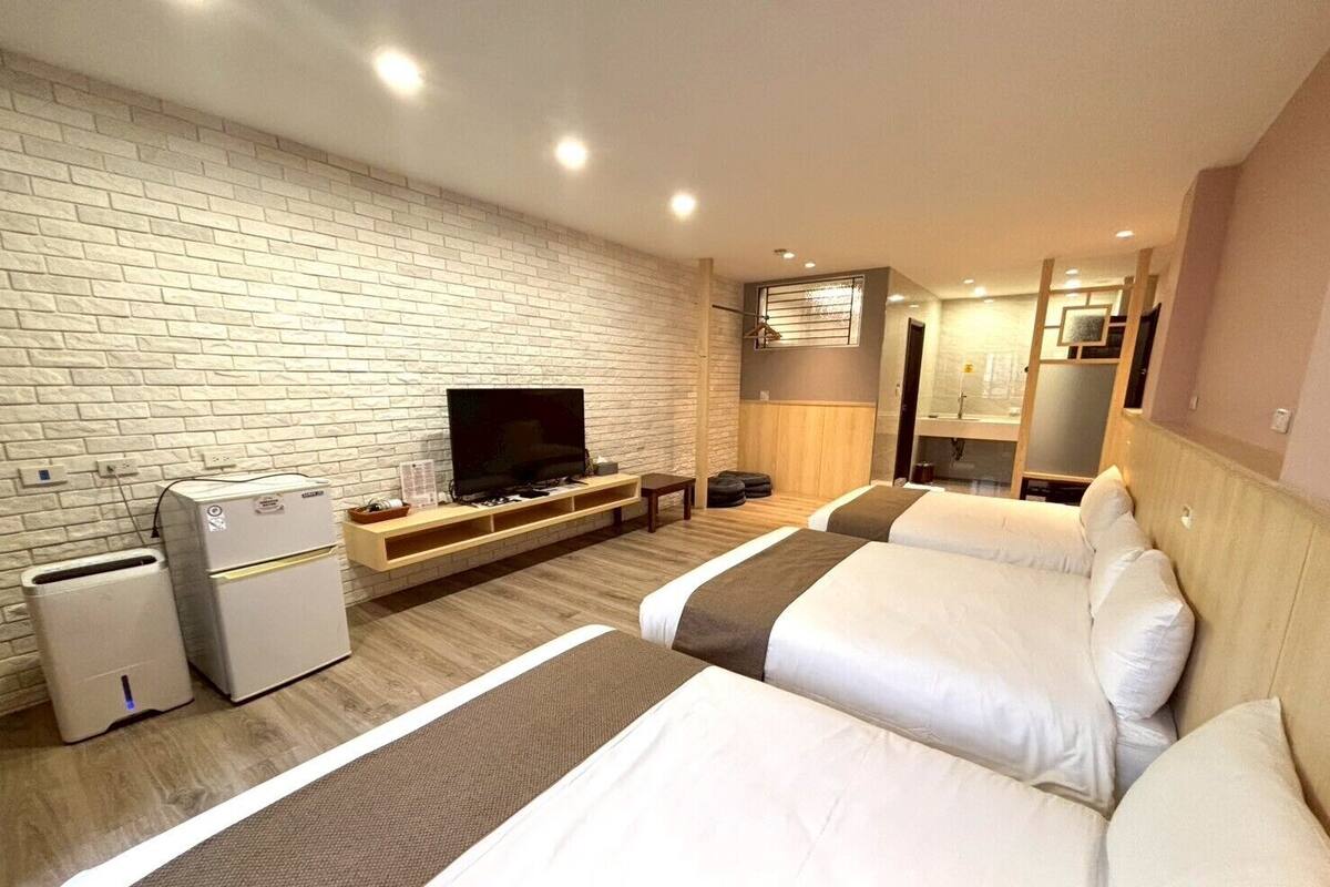 A hotel room with a brick wall, a flat-screen TV, a minibar, two beds, and a wooden headboard.