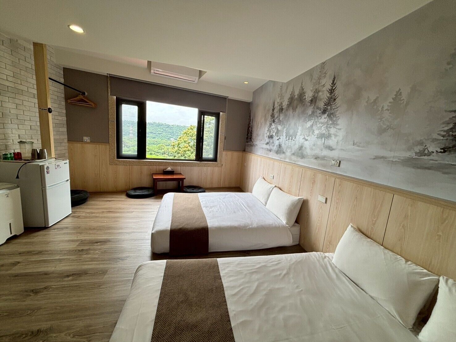 A modern hotel room with a large bed, a small desk, a refrigerator, and a window with a view of greenery.