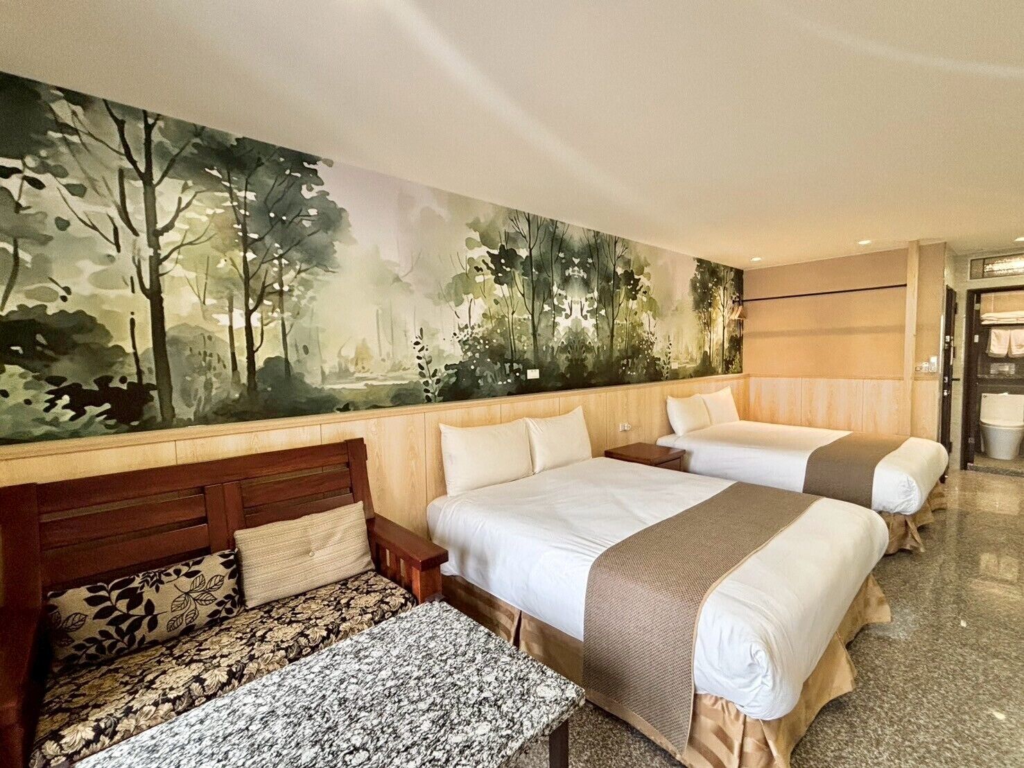 Une chambre d’hôtel avec deux lits, un banc et une grande fresque représentant une forêt sur le mur.