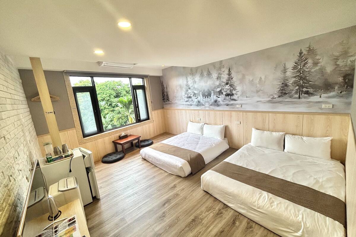 A modern bedroom with two beds, a wooden headboard, a large window with a view of trees, and a small table with a lamp.