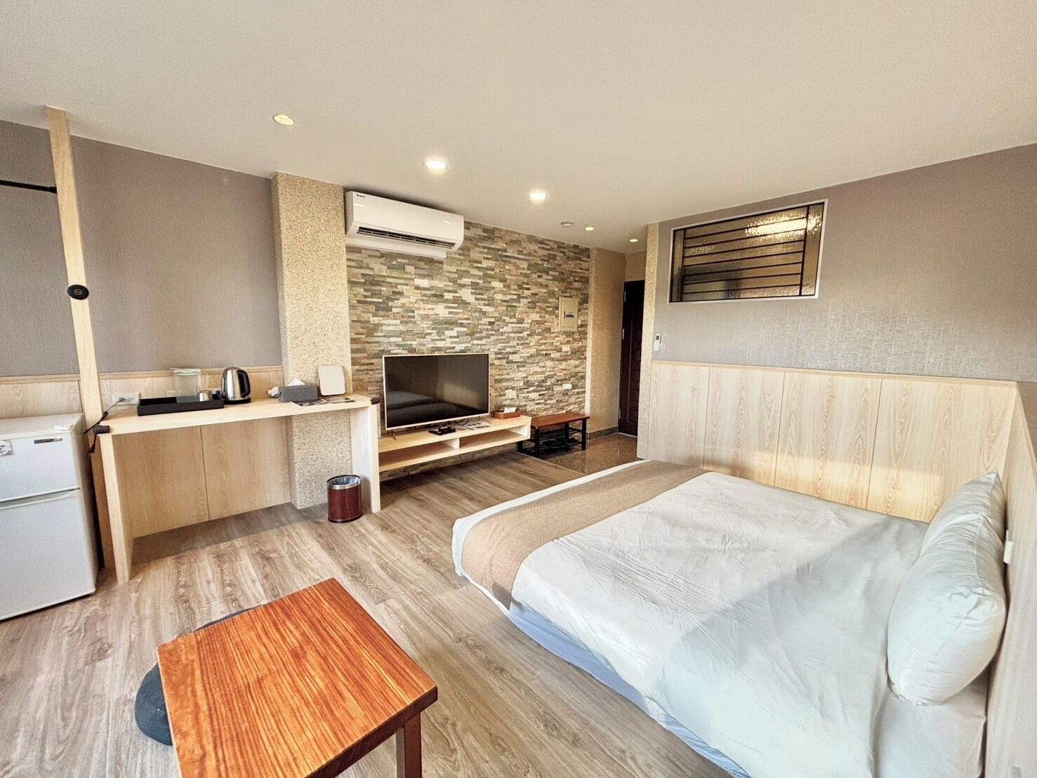 A modern hotel room with a wooden bed, a flat-screen TV, a small desk with a kettle, and a stone-patterned wall.