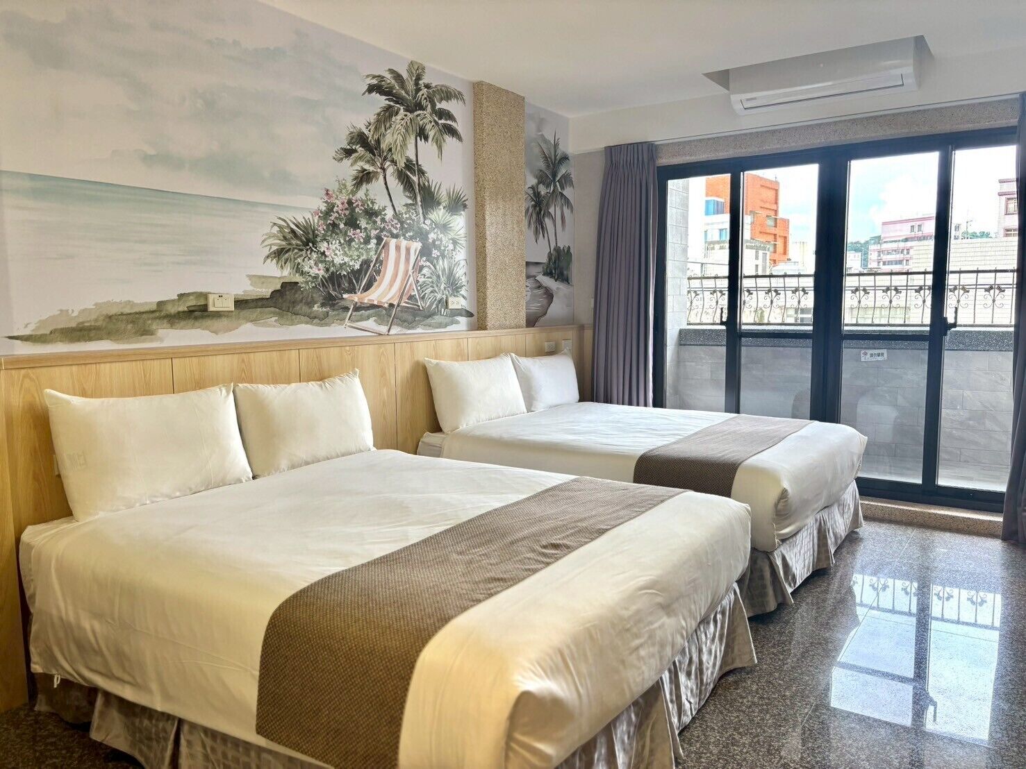Habitación de hotel con dos camas, un ventanal con vistas a edificios y un mural que representa una playa.