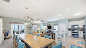 Une cuisine spacieuse équipée d’appareils modernes, un coin repas avec une grande table en bois et des chaises bleues, et une vue sur l’océan par de grandes fenêtres.