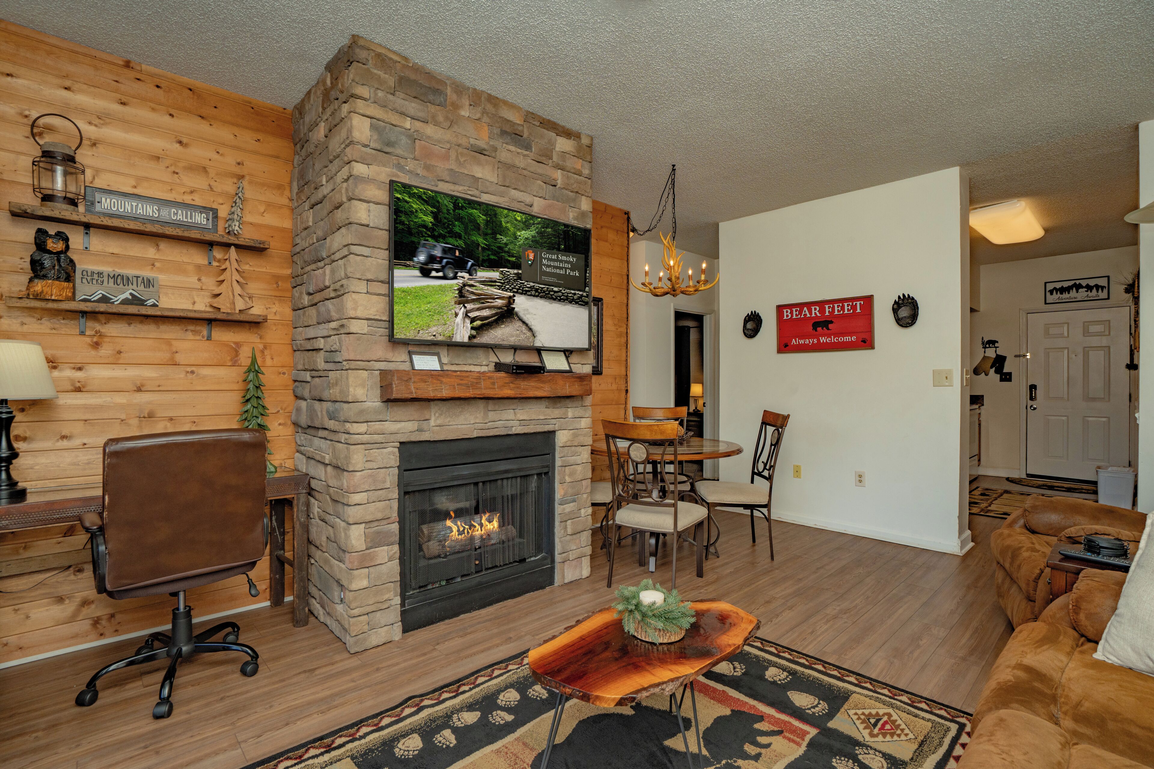 A living room with a stone fireplace, wooden walls, a television mounted on the fireplace, and a sign reading 'Great Smoky Mountains National Park'.