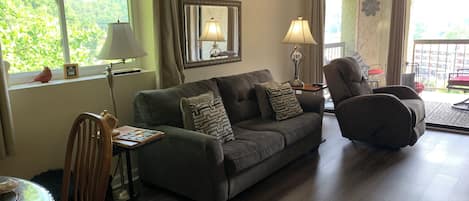 Una sala de estar con sofá, sillón y mesa de comedor. Se ve un ventanal con vistas a la vegetación.