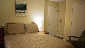 Un dormitorio con cama, una silla, una mesita de noche, una lámpara y una puerta.