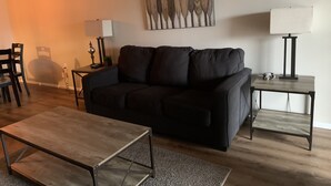 Una sala moderna con un sofá negro, una mesa de centro de madera, un cuadro en la pared y una lámpara sobre una mesita auxiliar.