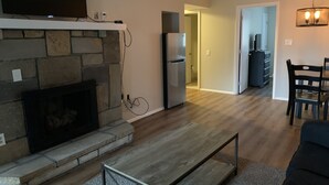 Una sala de estar con chimenea de piedra, un comedor con mesa y sillas, una cocina con refrigerador y un pasillo que conduce a otra habitación.
