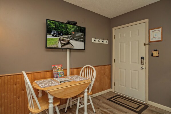 A dining area with a wooden table and chairs, a television mounted on the wall, and a door with a coat hook.
