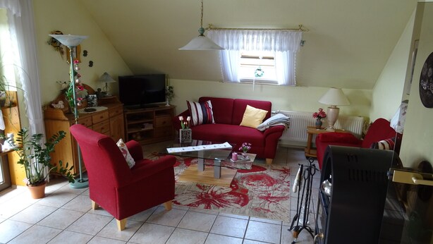 A cozy living room with a red sofa, a glass coffee table, and a television. There is a window with curtains, a radiator, and a lamp. - Apartment house Monika (Bergenhausen)